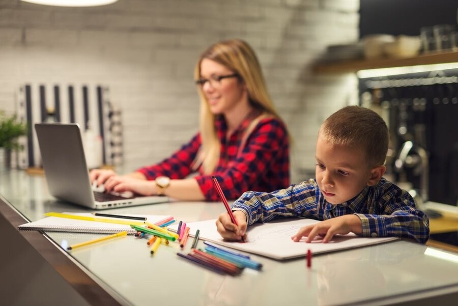 A mom working at her computer at a kitchen counter with her son coloring next to her.
