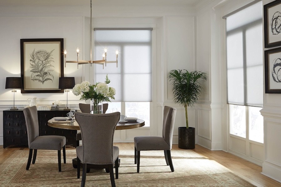 A brightly lit dining room with large windows covered almost entirely with motorized shades.